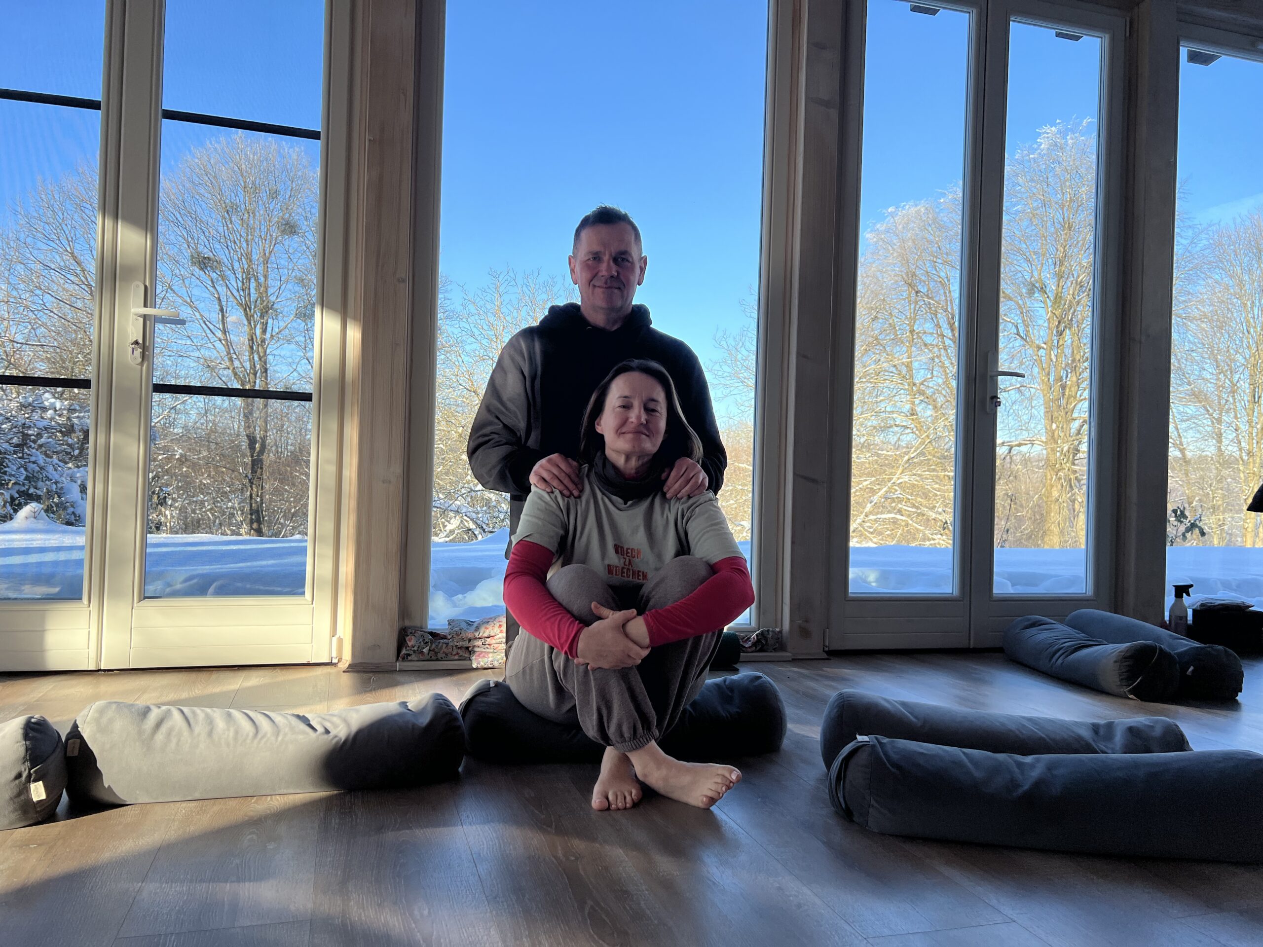 Mężczyzna i kobieta, uśmiechnięci, znajdują się w jasnym pokoju do jogi z dużymi oknami wychodzącymi na zaśnieżony, słoneczny krajobraz. Kobieta siedzi boso w swobodnej pozie na poduszce do jogi, a mężczyzna stoi za nią, delikatnie trzymając dłonie na jej ramionach. Na drewnianej podłodze leżą porozrzucane wałki do jogi. Zdjęcie emanuje spokojem i wsparciem.