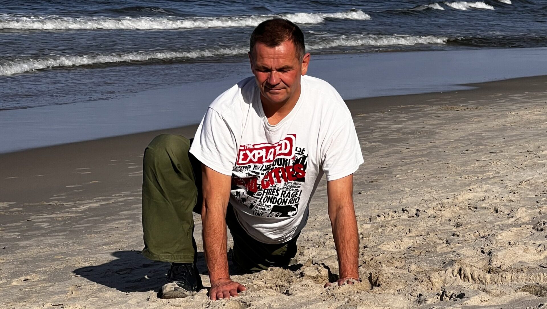 Mężczyzna wykonuje na plaży pozycję niskiego wykroku (Anjaneyasana), idealną dla biegaczy na rozciągnięcie bioder. Ma na sobie biały T-shirt z nadrukiem i zielone spodnie. Ćwiczy na piasku przy brzegu morza, w tle widać fale i niebieskie niebo z małym żaglowcem w oddali.