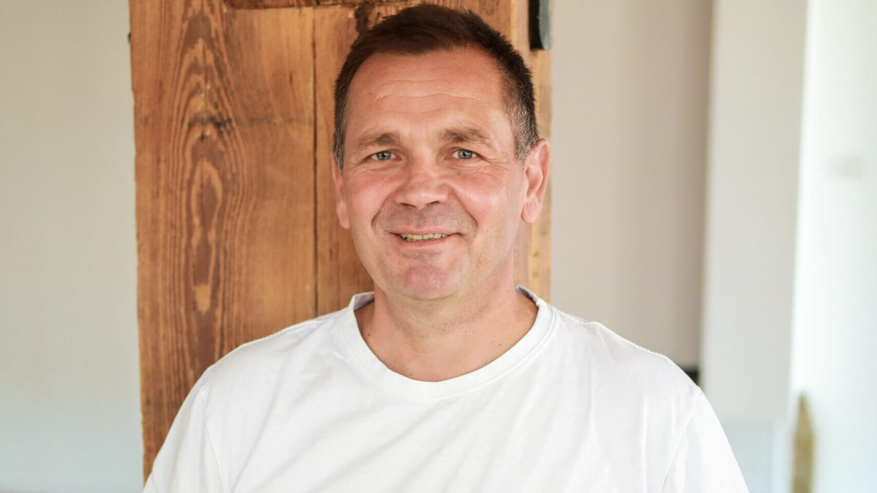 Grzegorz Michałowski, trener oddechu, uśmiecha się, stojąc w jasnym pomieszczeniu. Ma na sobie biały t-shirt z logo „Centrum Ciało Umysł Emocje”
